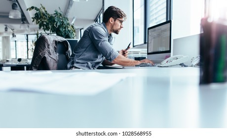 Young man sitting in office and working on desktop pc. Businessman looking at computer monitor while working in office. - Shutterstock ID 1028568955