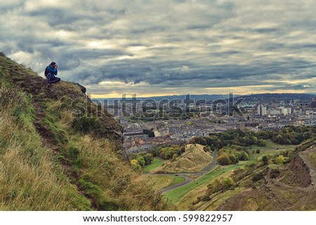 Young man sitting on Arthur's Seat, photographing the city of Edinburgh, Scotland.
Foreground of Salisbury crags, and in the backgrounds, houses and roofs with Edinburgh castle.