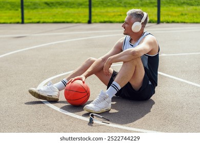 Young Man Sitting on Basketball Court Enjoying Music While Resting Next to a Basketball During a Sunny Day - Powered by Shutterstock