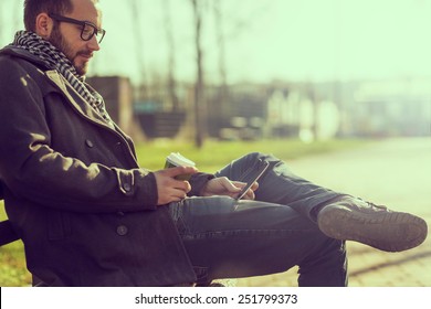 Young man sitting on a bench, reading an e-book on a tablet and drinking coffee  - Powered by Shutterstock