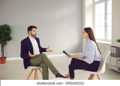 Young Man Sitting On Chair In Television Studio And Giving Interview To Journalist. Happy Smiling TV Host With Microphone Talking To Famous Person And Asking Questions About His Opinion And Experience