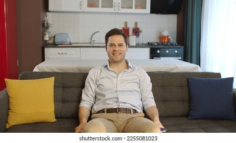 Young man sitting on comfortable sofa in his apartment and looking at camera with smile. Creative people can put whatever they want where the man points. - Shutterstock ID 2255081023