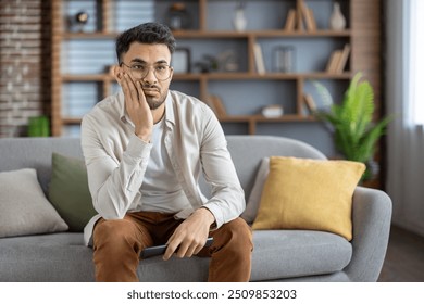 Young man sitting on comfortable sofa holding remote control, appearing bored and disengaged with television content. Casual clothing, glasses, and home setting convey relaxation and lack interest.