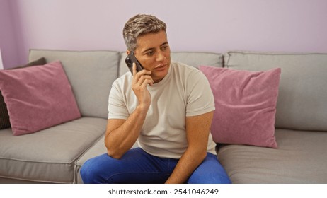 Young man sitting on a couch in a living room talking on his phone, with pink and grey cushions, wearing a white t-shirt and blue jeans, with a focused expression - Powered by Shutterstock