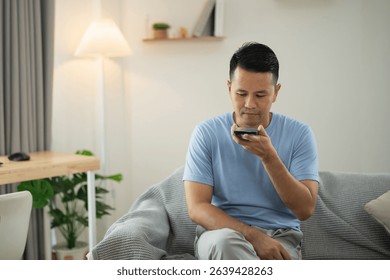Young man sitting on a couch in a modern living room, using smartphone voice assistant for hands-free technology and convenience in daily tasks - Powered by Shutterstock - Get 15% off with code: PIKWIZARD15