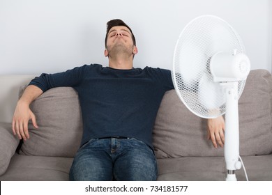 Young Man Sitting On Couch Cooling Off With Fan During Hot Weather At Home