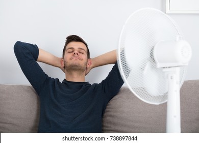 Young Man Sitting On Couch Cooling Off With Fan During Hot Weather At Home