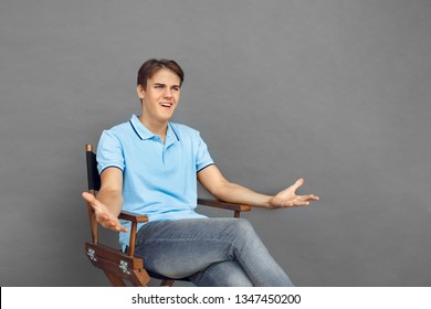 Young Man Sitting On Director's Chair Isolated On Grey Wall Hands Aside Looking At Scene Irritated