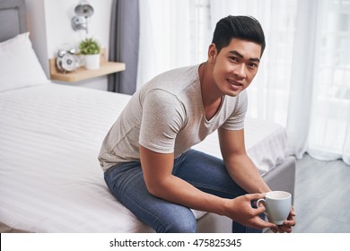 Young Man Sitting On Edge Of Bed And Drinking Coffee