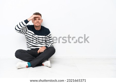 Young man sitting on the floor isolated on white background looking far away with hand to look something