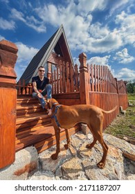 Young Man Sitting On Front Porch Of Wooden House With His Dog. Male Playing With Hungarian Vizsla Pointer Near Tiny Wooden House In Sunny Summer Morning. Man Traveler With Pet Resting In Countryside.