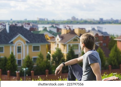 Young man sitting on the grass dreaming about his own house. Urban landscape with village 