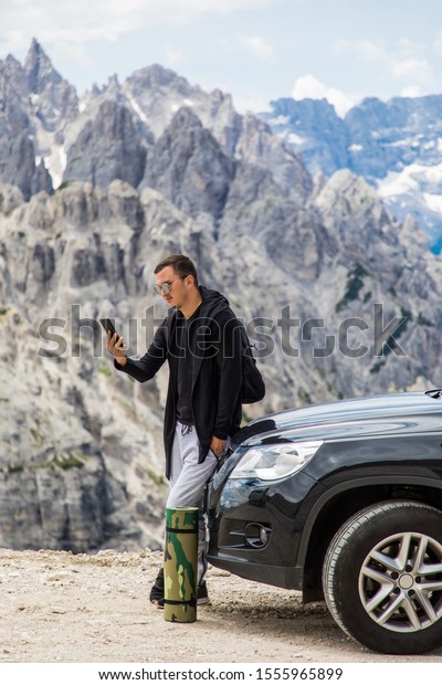 Young man\
sitting on hired car and talking on the mobile phone on mountains\
background. Car hire. Car\
trip.