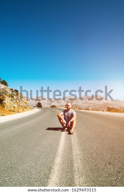 A young man is sitting on a mountain\
road. The dividing line on the road goes into the horizon. Mountain\
landscapes of the island of Crete. Travel by\
car