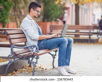 Young Man Sitting On The Park Bench With Laptop On His Lap