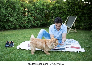 Young Man Sitting On Picnic Blanket On The Grass In Backyard And Feeding His Cute Pet Dog	