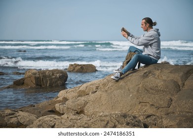 Young man sitting on a rocky shore, using his smartphone to capture the scenic ocean view. Wearing casual clothing and a hoodie, enjoying a peaceful moment by the beach. - Powered by Shutterstock