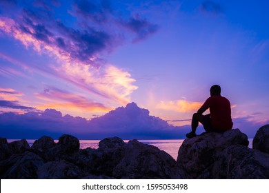 Young man sitting on sea rocks by the beach thinking, contemplating, determining the way forward. Life changing decisions. Priority decision making. Freedom to choose. Passing time as the sun sets.