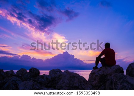 Young man sitting on sea rocks by the beach thinking, contemplating, determining the way forward. Life changing decisions. Priority decision making. Freedom to choose. Passing time as the sun sets.