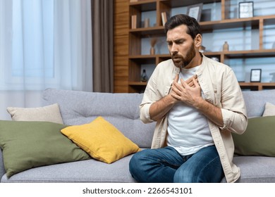 A young man is sitting on the sofa at home and holding his chest. Feels severe pain, shortness of breath, panic attack, heart attack, stroke.