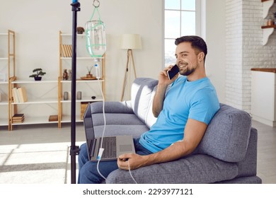 Young man sitting on sofa and talking by mobile phone at home with a laptop while receiving IV drip infusion and vitamin therapy in his blood. Male person receiving injection therapy.
