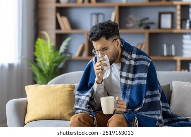 Young man sitting on sofa covered with blanket, holding hot drink and tissue. He appears sick, blowing nose, wrapped in warm blanket, trying to feel better at home during illness or cold season. - Powered by Shutterstock