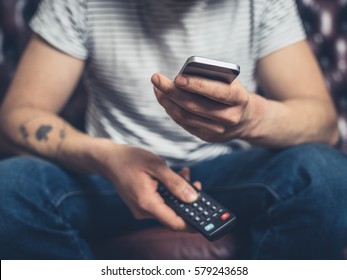 A young man is sitting on a sofa with a remote control and a smartphone