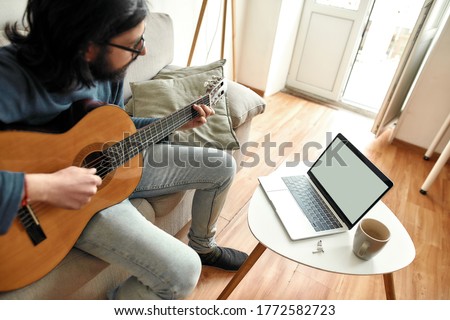 Young man sitting on sofa at home and learning how to play guitar, watching online course on laptop. Distance education. Main focus on laptop. Stay home. E-learning. Music school online