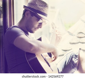 Young Man Sitting On The Window Playing The Guitar. Stylish Hipster Guy With Hat And Sunglasses Enjoys Music And Summer Holiday. Retro Filter.