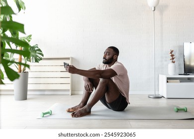 A young man sitting on a yoga mat at home, holding a smartphone, taking a break after exercising. The scene emphasizes wellness, relaxation, and modern fitness routines. - Powered by Shutterstock