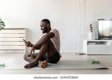 A young man sitting on a yoga mat at home, holding a smartphone, taking a break after exercising. The scene emphasizes wellness, relaxation, and modern fitness routines. - Powered by Shutterstock