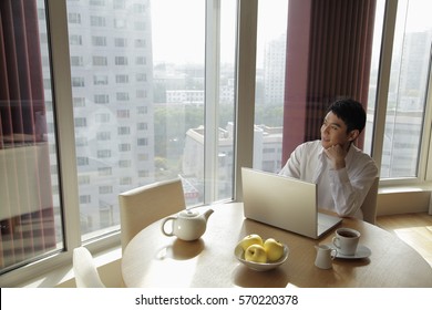 Young man sitting at a table with a computer looking out the window