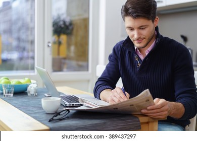 Young Man Sitting At A Table In His Kitchen At Home Searching The Classifieds In A Newspaper For Employment Or Accommodation