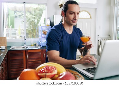 Young man sitting at the table typing on laptop computer and drinking orange juice in the kitchen - Shutterstock ID 1357188050