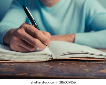 A Young Man Is Sitting At A Table And Is Writing In A Notebook