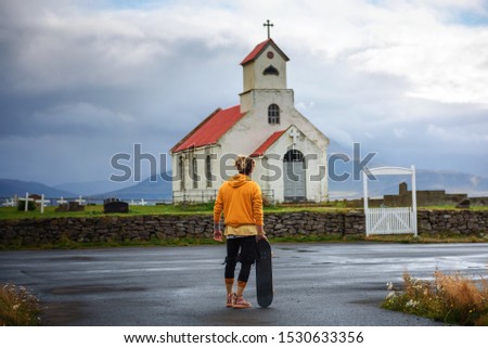 Young man with a skateboard looking at a church with a cemetery in Iceland. Loneliness, hope, sadness , depression and solitude concept.
