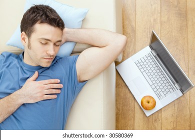 Young Man Sleeping On Sofa At Home, Laptop On Floor.  Overhead View.