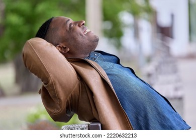 Young man with smile leaned back on back of bench in park with hands back behind his head, dreaming of the upcoming vacation. Cheerful black guy happily looks at the sky sitting on a bench - Powered by Shutterstock