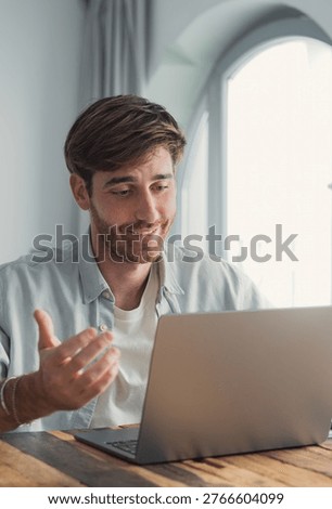 A young man smiles and gestures while on a laptop video call in a bright home office, conveying remote work, friendly communication, casual business meeting and modern digital collaboration.