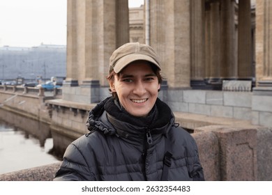 Young man smiles while standing on a bridge in a city with historic architecture during a cool day. waist up portrait