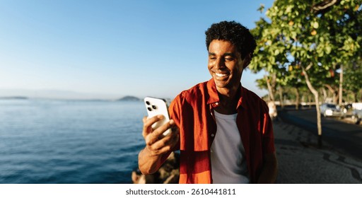A young man smiles while using his smartphone along Urca's Wall, with a scenic view of the ocean. The clear blue sky adds to the relaxed mood, capturing a perfect blend of technology and nature.