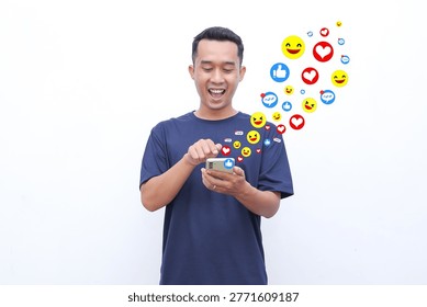 Young man smiles while using smartphone. Colorful emojis float above his head. He enjoys social media interaction. Image captures joyful digital engagement. Perfect for digital marketing visuals