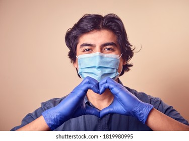 A Young Man Smiles While Wearing Nitrile Gloves With A Surgical Mask On His Face. Long Hair Man. Gloved Hands Making Heart Shape.