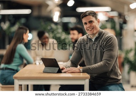 Young man smiling at camera while working on a digital tablet in a modern office with his colleagues. Teamwork and success concept
