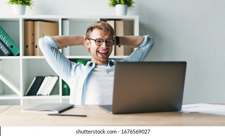 Young Man Smiling As He Reads The Screen Of A Laptop Computer While Relaxing Working On A Comfortable Place By The Wooden Table At Home. Happy Social Distancing