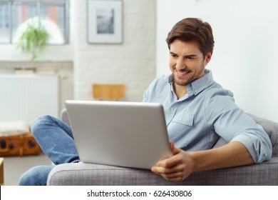 Young Man Smiling As He Reads The Screen Of A Laptop Computer While Relaxing On A Comfortable Couch At Home In His Jeans