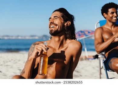 A young man smiling and holding a beer on a bright summer day at the beach. He is enjoying the relaxed atmosphere with friends, capturing the joy and warmth of summertime leisure. - Powered by Shutterstock