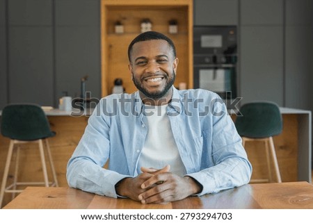 Young man smiling at a kitchen table, casually dressed for remote work or a video call. Warm home interior setting conveys friendliness, confidence and approachable lifestyle branding for portraits and business use.