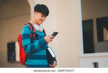 A young man is smiling and looking at his cell phone while holding a backpack and two books. He seems to be enjoying his time and is possibly checking his messages or browsing the internet - Powered by Shutterstock