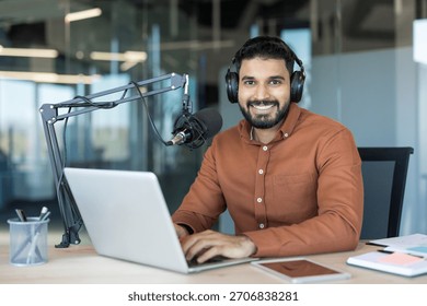 Young man smiling while broadcasting an online radio show or recording a podcast, speaking into a professional microphone and typing on a laptop in a modern office setup